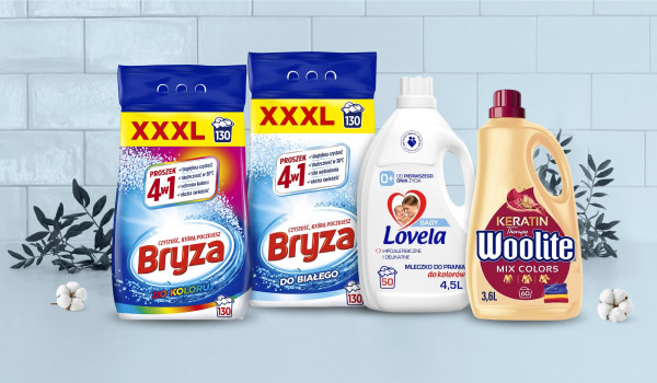 Cztery produkty do prania ustawione na tle jasnych płytek i dekoracyjnych gałązek. Widoczne są opakowania proszków Bryza z oznaczeniem XXXL, mleczko do prania Lovela Baby o pojemności 4,5 litra oraz płyn Woolite Keratin Mix Colors o pojemności 3,6 litra.