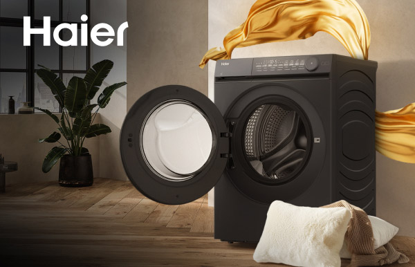 Na zdjęciu znajduje się czarna pralka z otwartymi drzwiami, ustawiona na drewnianej podłodze, z widocznym wnętrzem bębna. W tle widać roślinę w doniczce oraz złotą tkaninę unoszącą się w powietrzu, a w lewym górnym rogu znajduje się logo Haier.