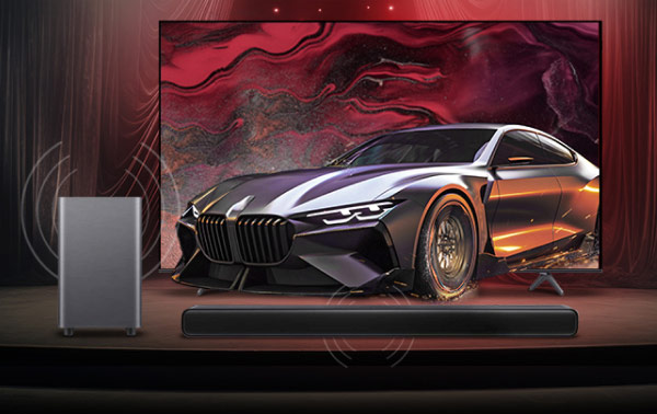 Na zdjęciu znajduje się duży telewizor z wyświetlonym obrazem sportowego samochodu na tle abstrakcyjnej, czerwono-czarnej grafiki. Przed telewizorem umieszczony jest soundbar oraz subwoofer.